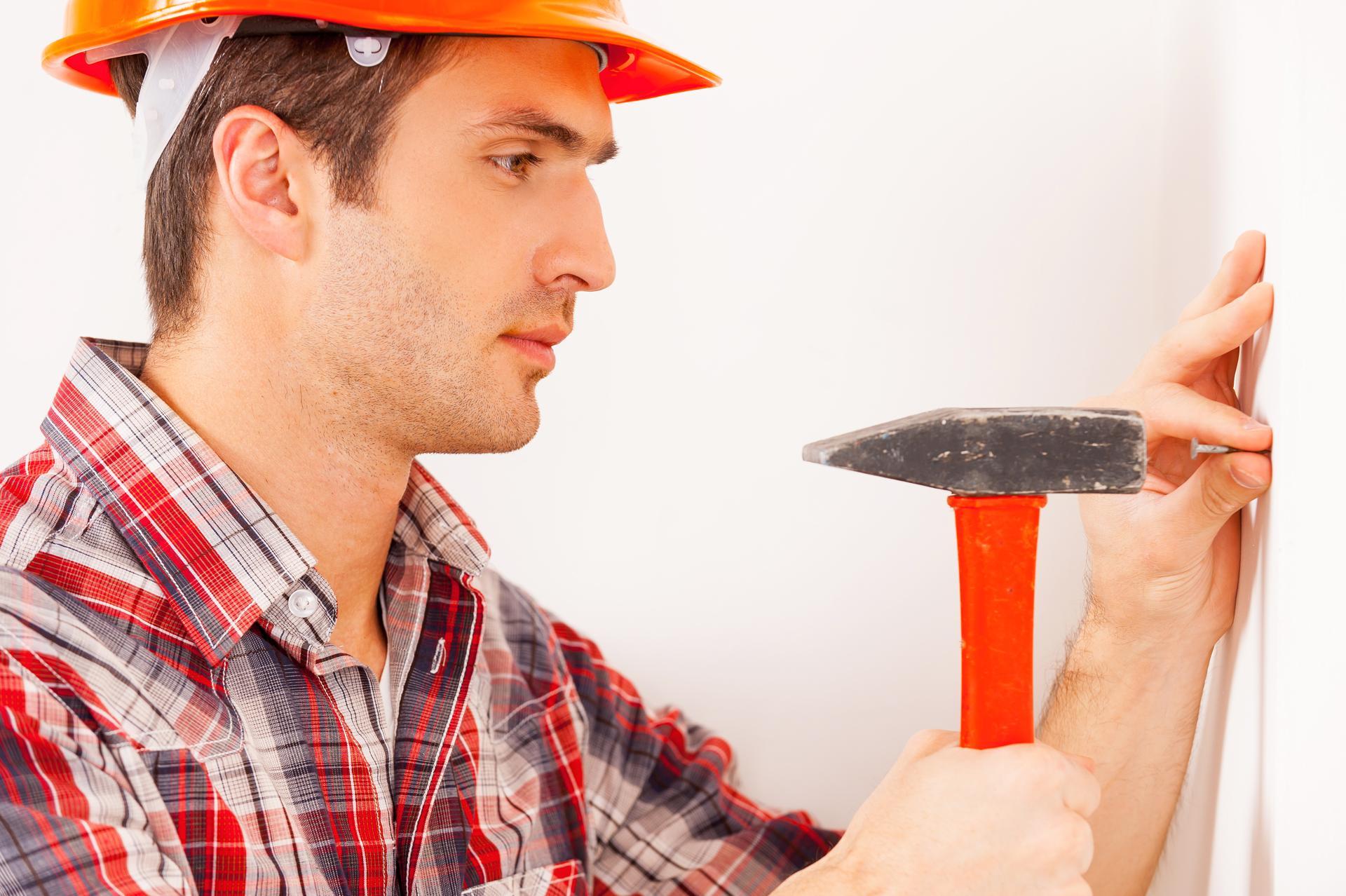 Home improvement. Side view of handsome young handyman in hardhat hammering a nail while standing near the wall