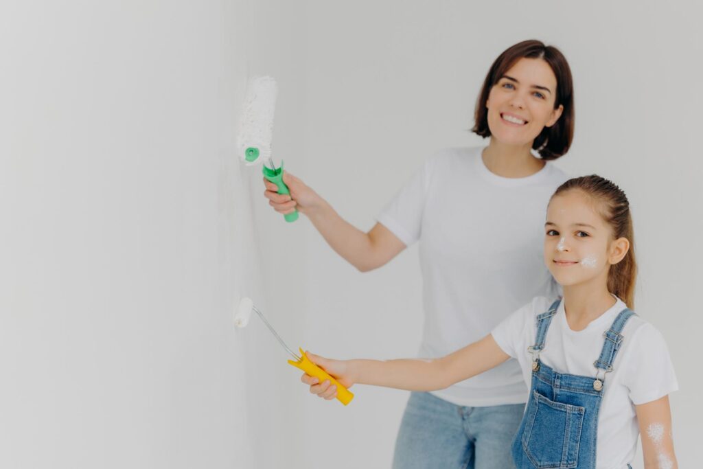 Horizontal shot of happy girl and her mother paint walls in white color, use paint rollers, improve house, busy with domestic work, move in new apartment. Home repair and improvement concept.